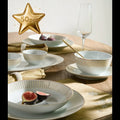 A 50-piece white ceramic tableware set with gold-toned rim detailing, styled on a wooden dining table with gold napkins, plates, bowls, a wine glass, and figs. A gold star graphic with '50pcs' text is in the top left corner.
