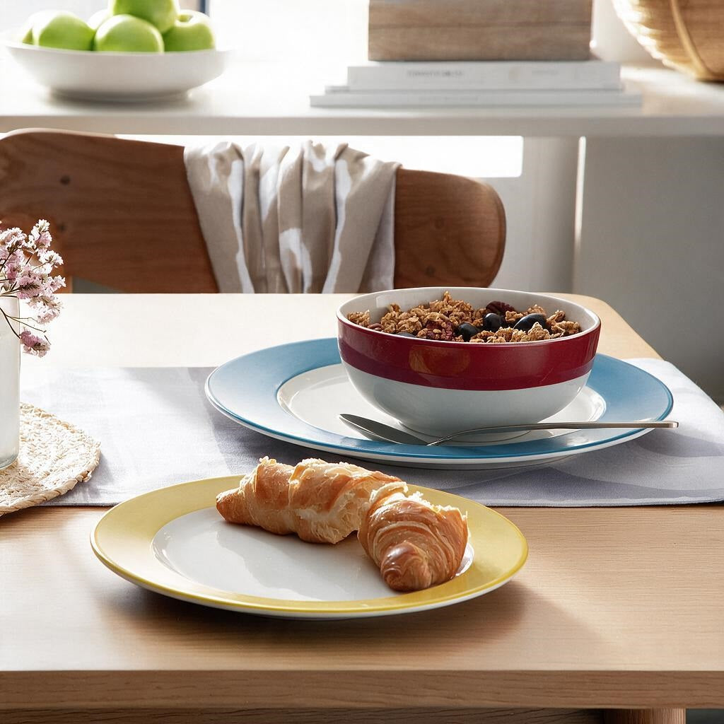 A breakfast table setting with two ceramic plates, a red and white bowl of granola, and a croissant on a light wood table in a bright kitchen setting.