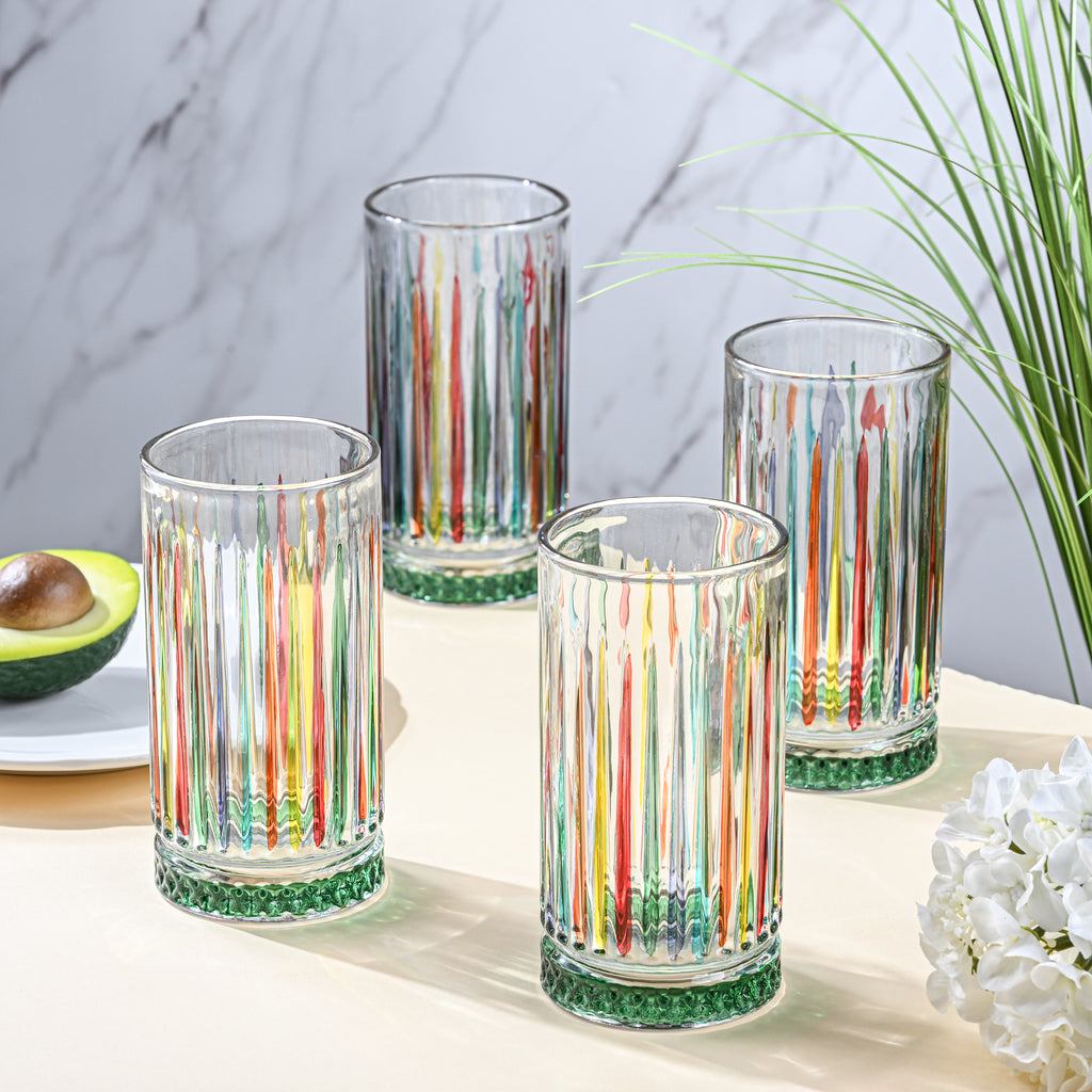 Set of four colorful striped glasses on a table with a plant and avocado in the background.